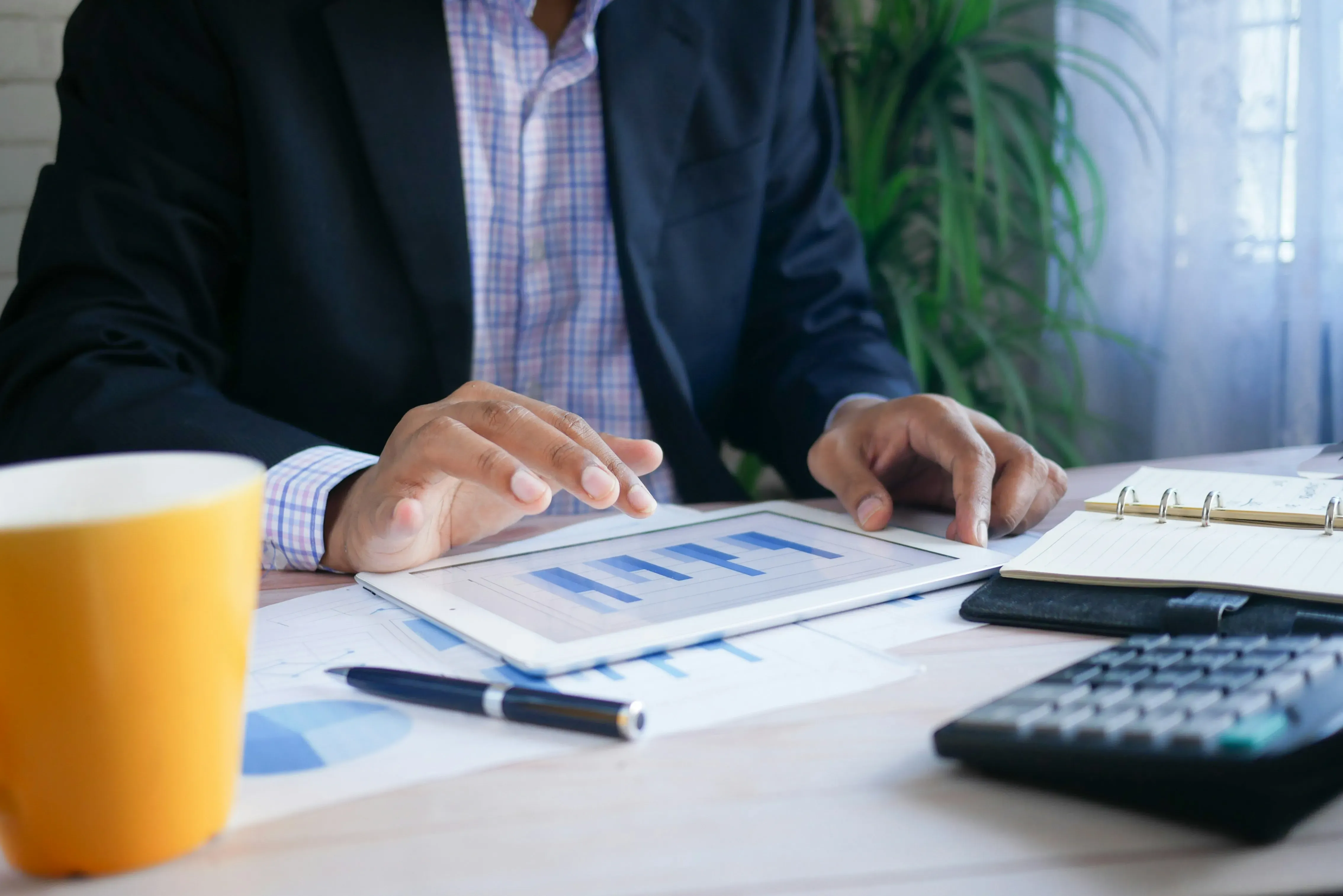 Person in business attire analyzing bar charts on a tablet with a pen, notebook, and calculator on the desk.