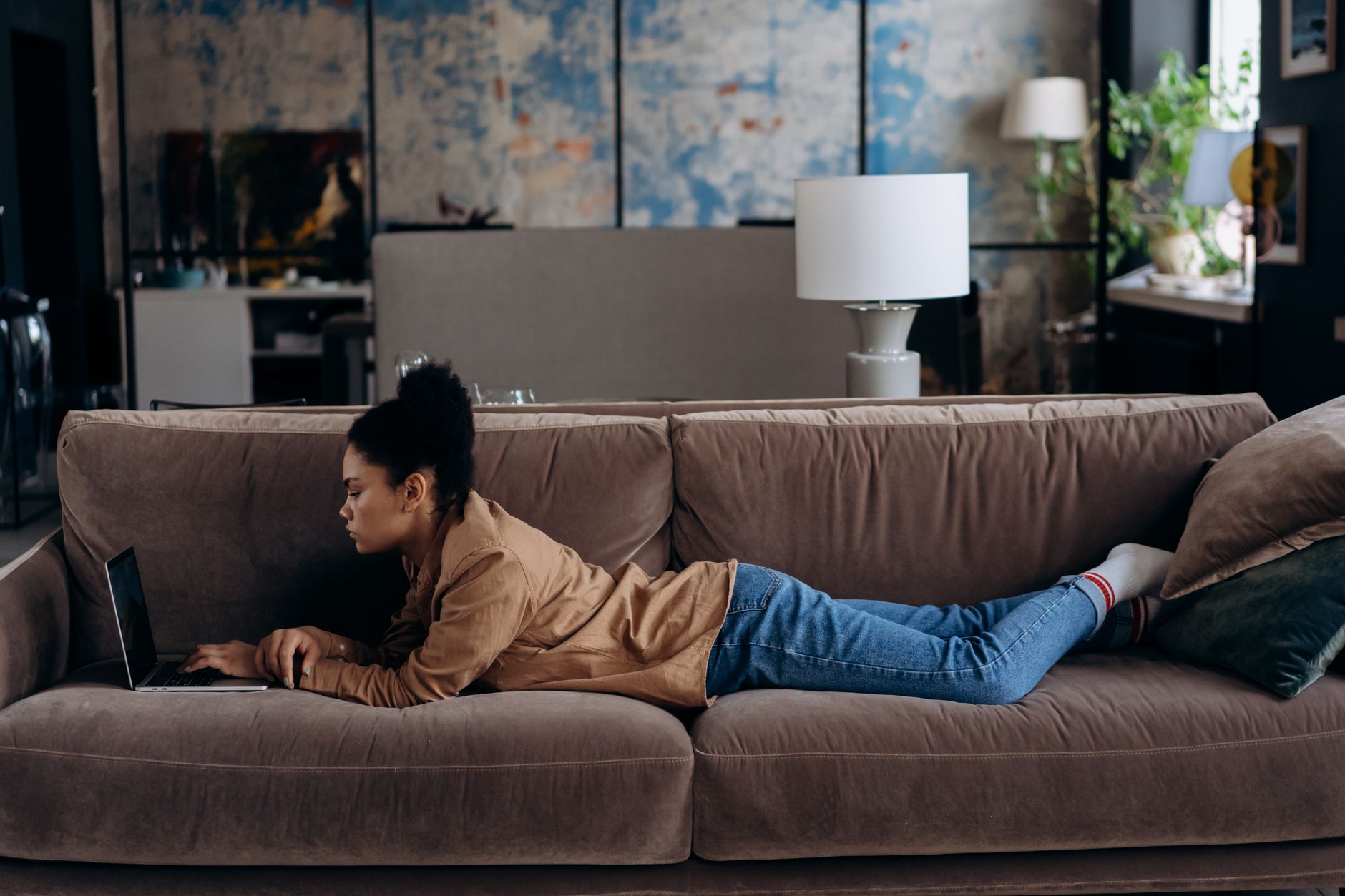 African American woman lying on sofa using laptop for remote work at home.