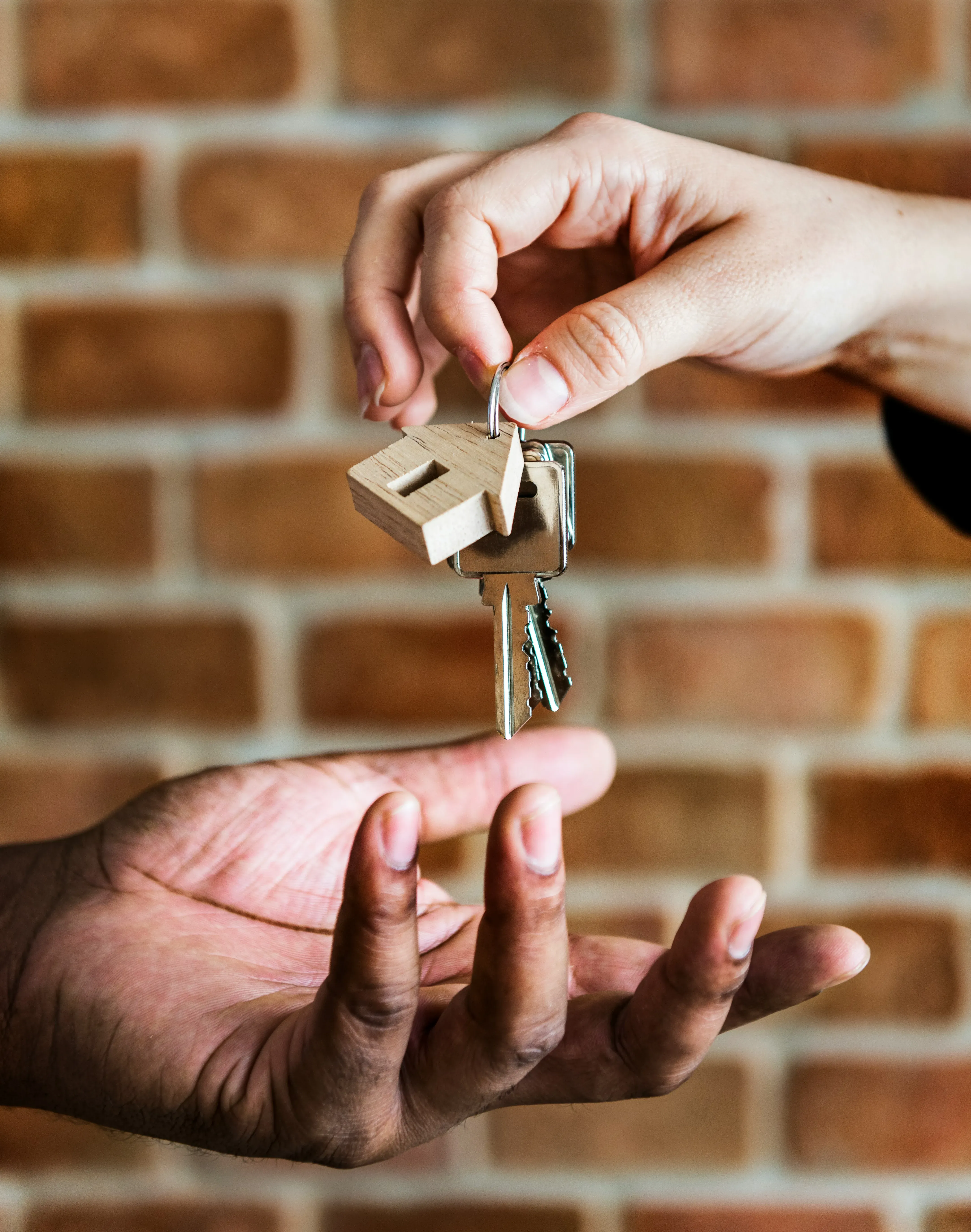 Close-up of one hand handing over house keys with a wooden house-shaped keychain to another outstretched hand against a brick wall background.