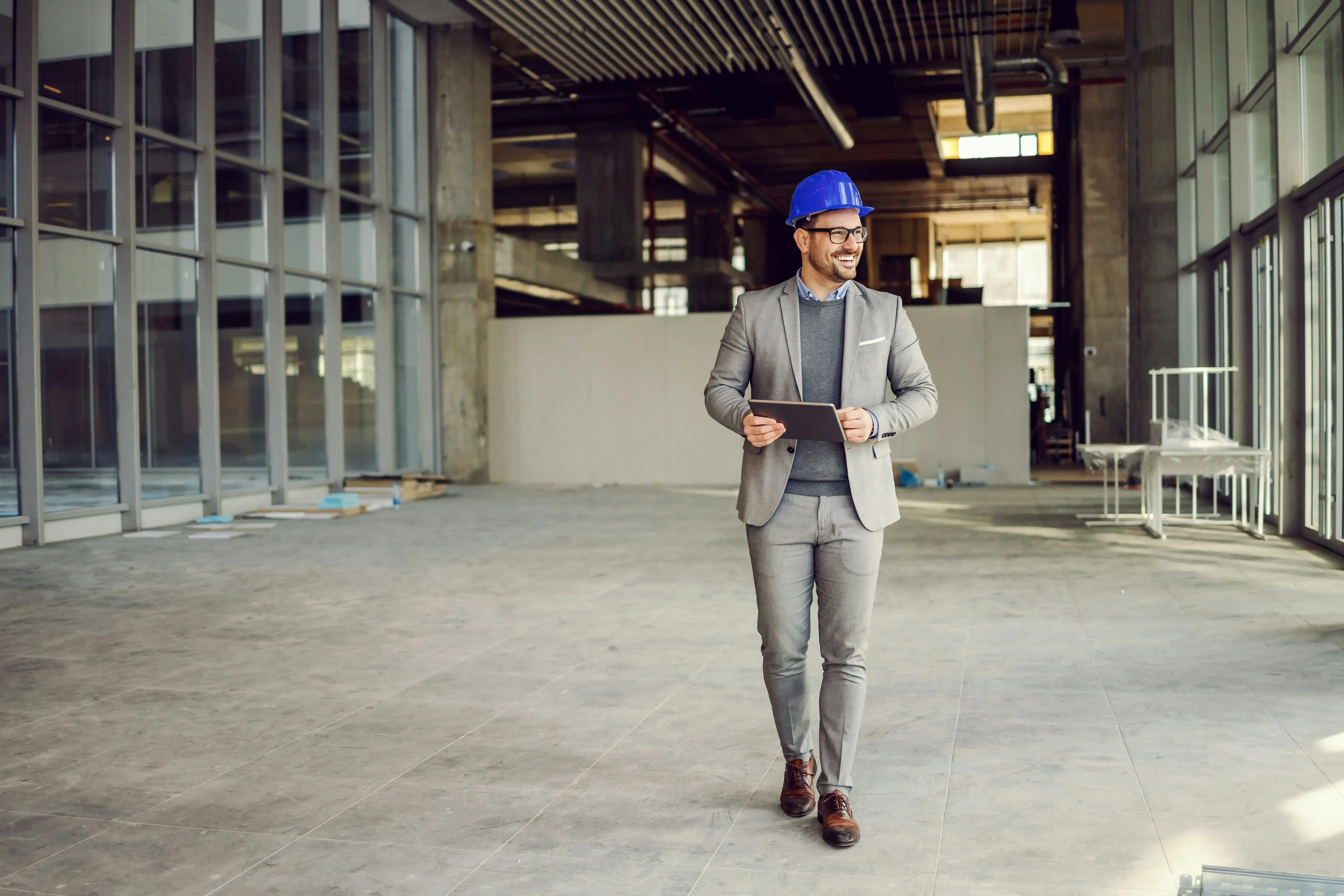 Man in a grey suit and blue hard hat holding a tablet walking inside an empty commercial building under construction.