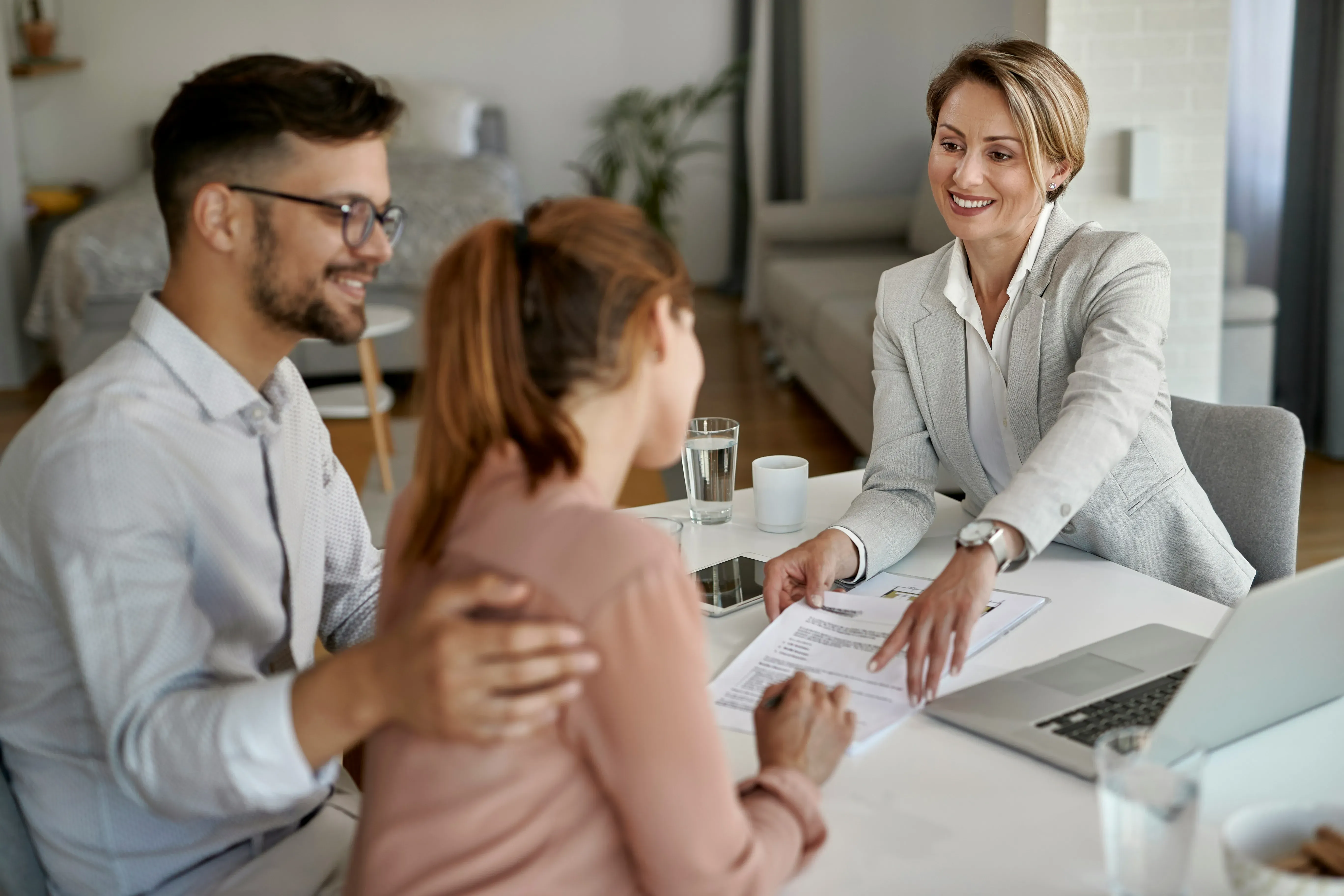 Smiling real estate agent showing a contract to a couple sitting at a table with a laptop and glasses of water.