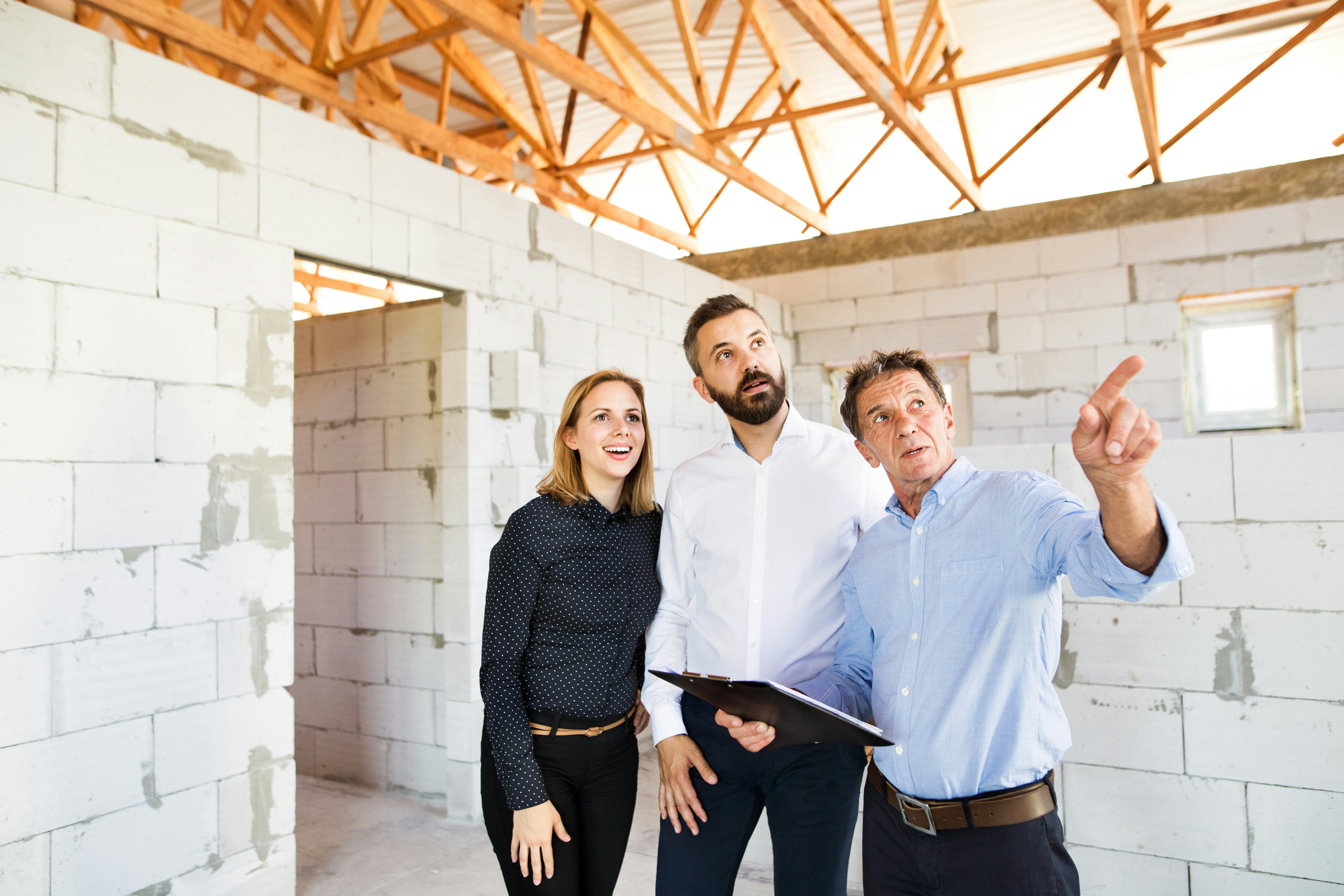Three people inside a partially built house, with one man pointing and holding a clipboard while the others look on attentively.