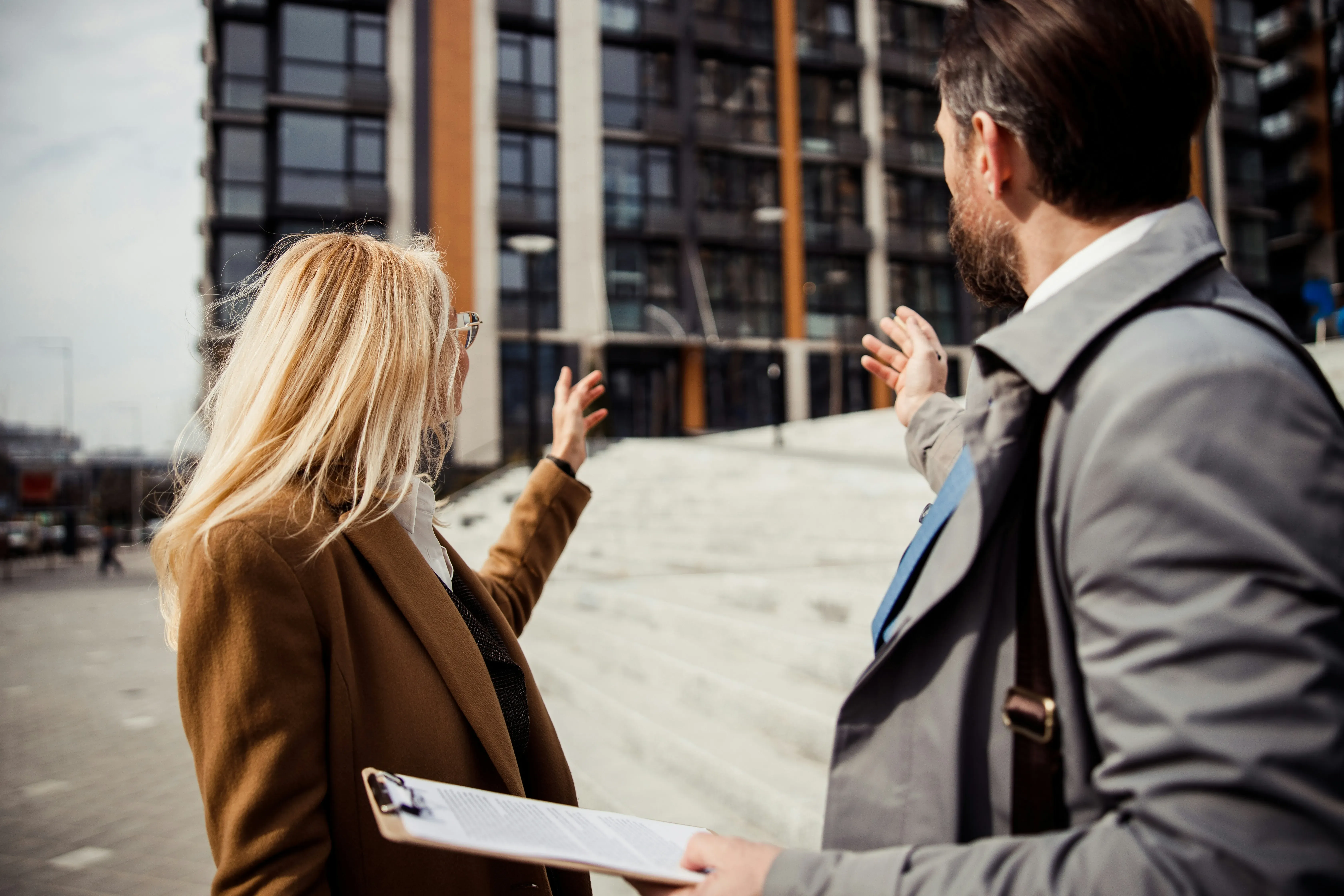 Two professionals, a woman with a clipboard and a man in a trench coat, gesturing towards a modern building.
